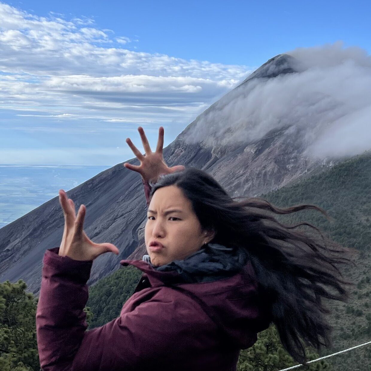 Grace standing in front of a smoking active volcano. She is raising both hands to mimic an explosion and making a funny face at the camera.